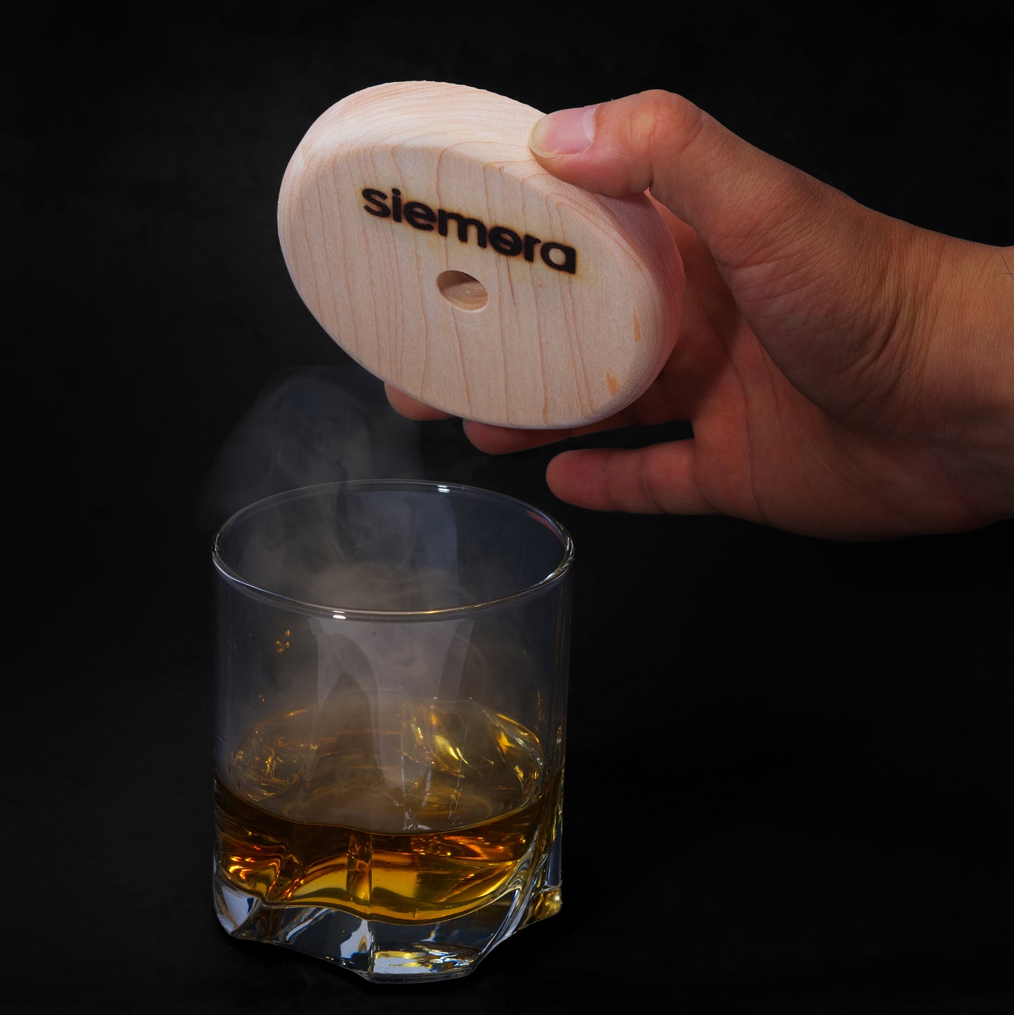 Hand holding a wooden cocktail smoker labelled 'siemora' underneath it over a glass of whiskey being, displayed on a black background