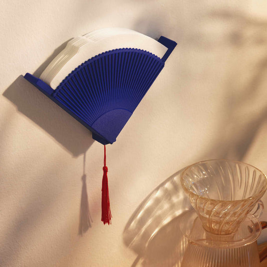 Blue coffee filter holder with a red tassel mounted on the white wall  next to a glass V60 dripper