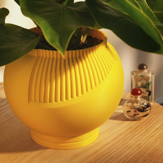 3D Printed yellow planter pot with a green leaf plant in it sitting on a wood table next to decorative glass bottles
